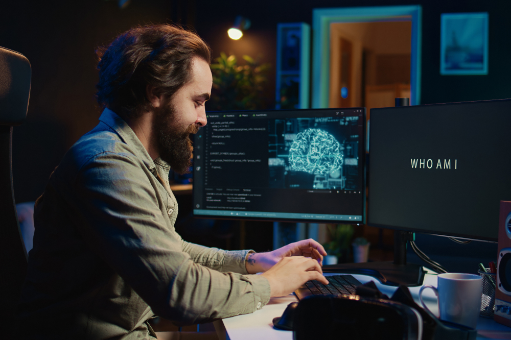 A bearded man works on AI programming at a desk with dual monitors, one showing a neural network graphic and the other displaying the text "WHO AM I."