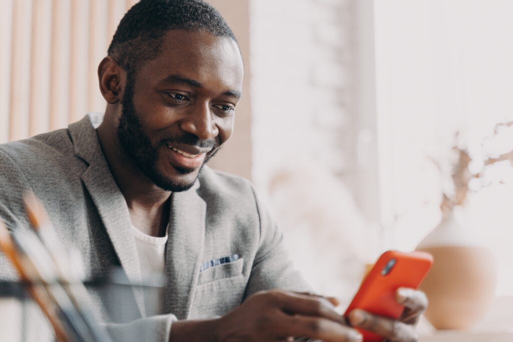 Smiling businessman in a grey blazer using a red smartphone while seated at a desk in a bright, modern office.
