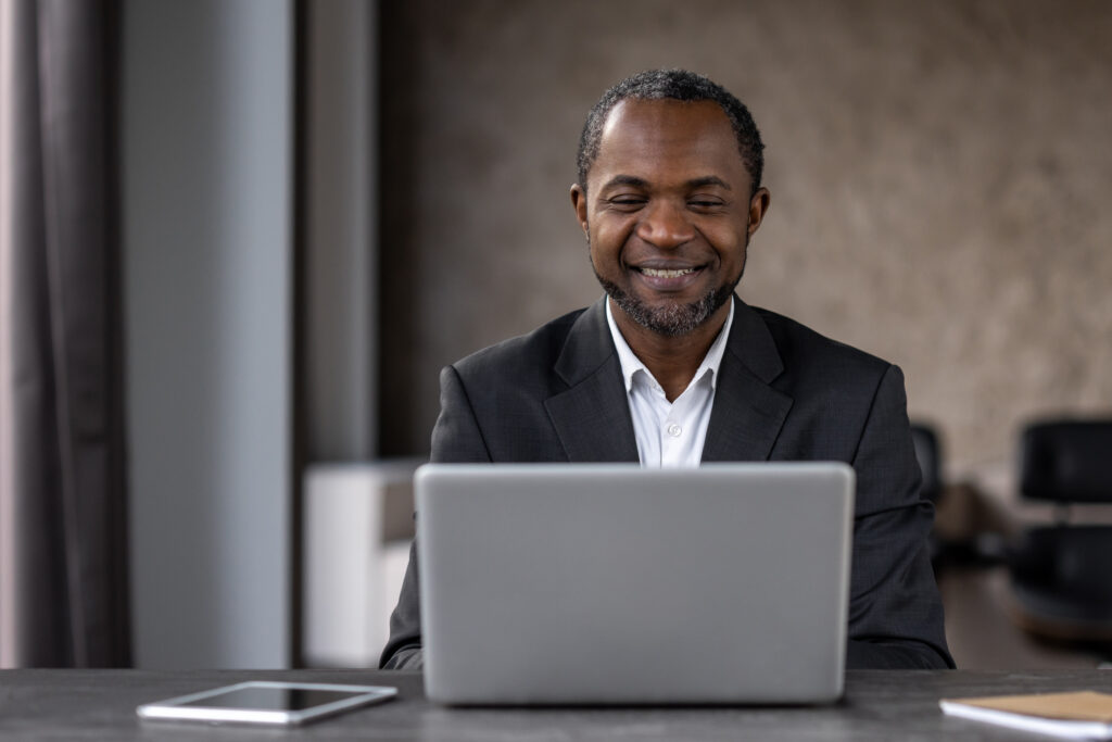 A smiling Black man in a dark suit and white shirt working on a silver laptop at a desk in a professional office setting.