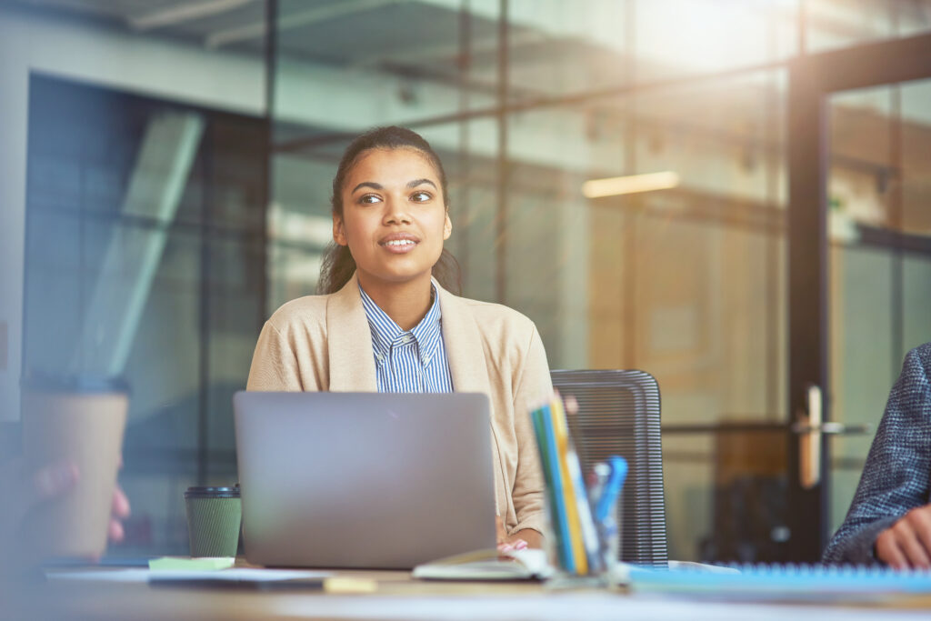 A young woman with dark hair tied back, wearing a beige cardigan and blue striped shirt, looking thoughtfully to the side while sitting at a laptop in a bright office.