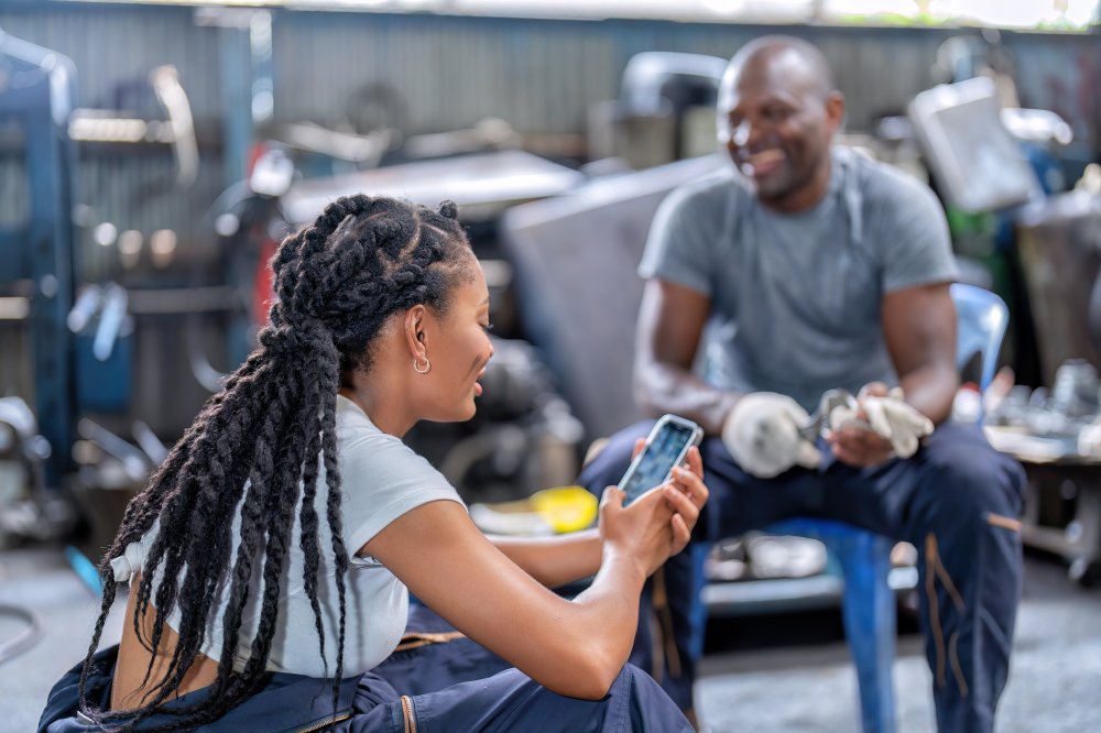 A female mechanic with braided hair sits in a garage, smiling as she shows something on her smartphone to a male colleague who is sitting across from her holding a wrench.