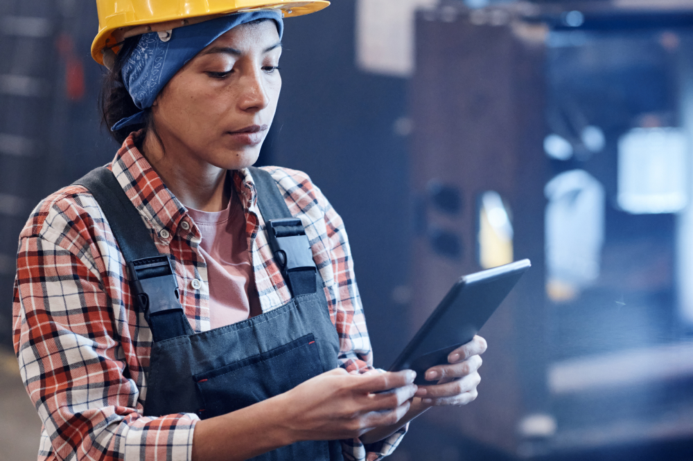 A focused female worker wearing a yellow hard hat, blue bandana, and denim overalls stands in an industrial plant while using a digital tablet to access work information.