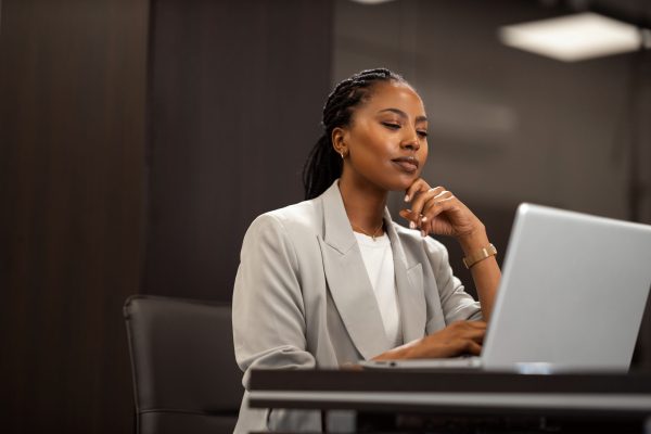 A woman with braided hair wearing a grey blazer sits at a desk, looking thoughtfully at her laptop screen.