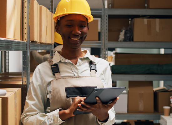 A smiling warehouse worker in a yellow hard hat using a digital tablet for real-time engagement.